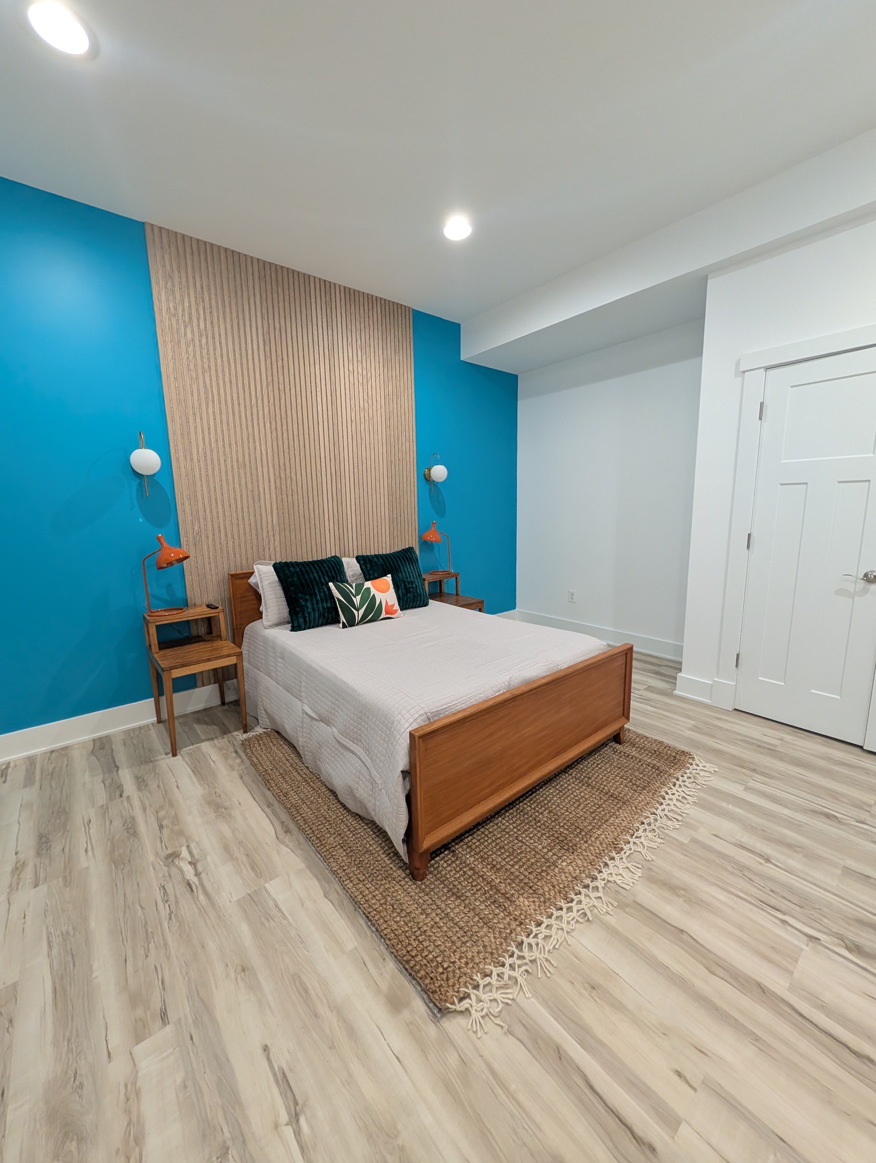 Finished basement bedroom with a teal accent wall, wood slat panel behind the headboard, and matching orange lamps on wood nightstands