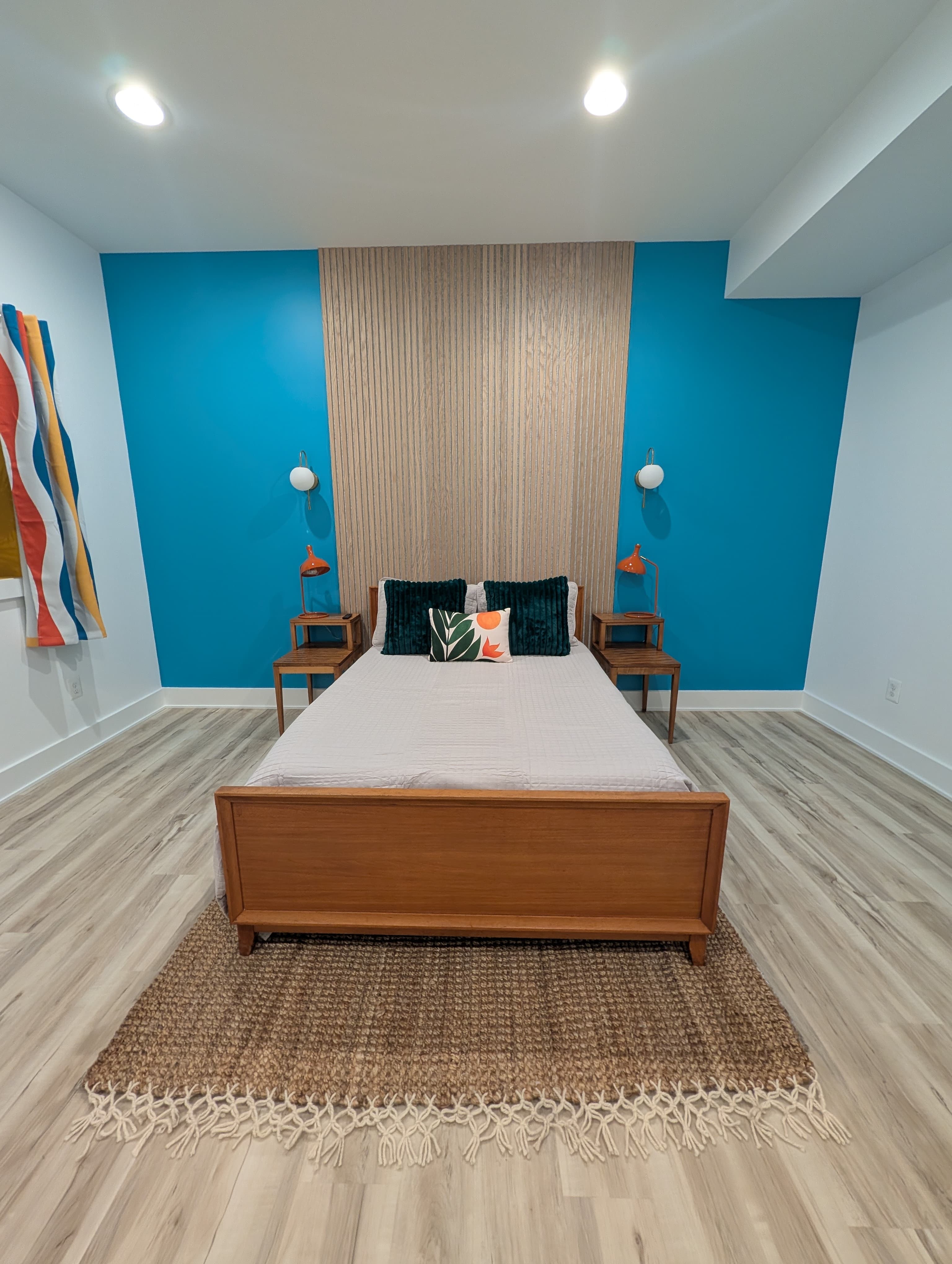 Finished basement bedroom, front view of the bed between two nightstands with orange lamps, facing the wood slat accent wall